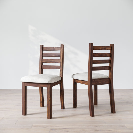 Two wooden dining chairs with a natural finish and upholstered seats, with a ladder back design, placed side by side on a bright floor against a white wall.
