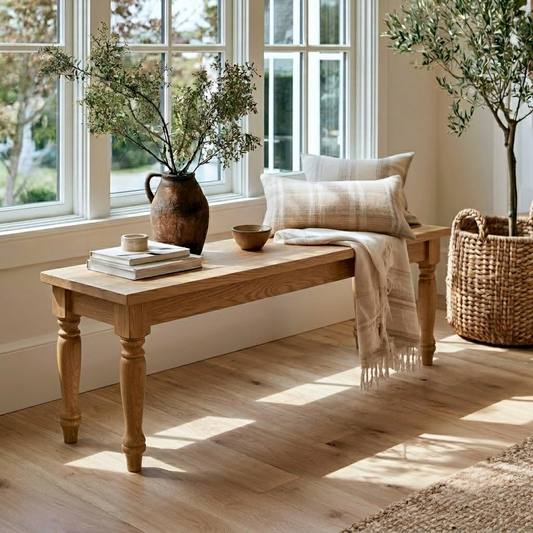 A rustic wooden bench with turned legs sits by a bright window in a sunlit room. The bench is styled with a dark brown ceramic vase of greenery, stacked books, a small bowl, and a beige plaid throw pillow with a cream fringed throw blanket draped over it.