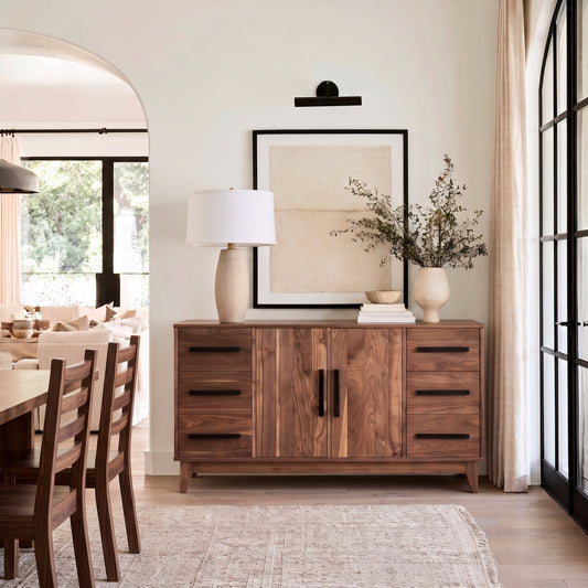 A mid-century modern wooden sideboard cabinet in a warm walnut finish, styled in a dining room with a table, chairs, a lamp, and vases.