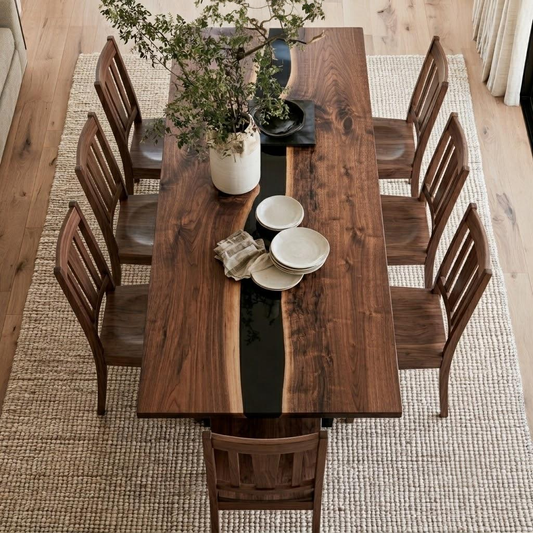 A rectangular wooden dining table with a live edge and a glossy black inlay, surrounded by six matching wooden chairs. The table is styled with a white ceramic vase of green branches, a black tray, and white plates on a woven beige rug over light wood flooring.