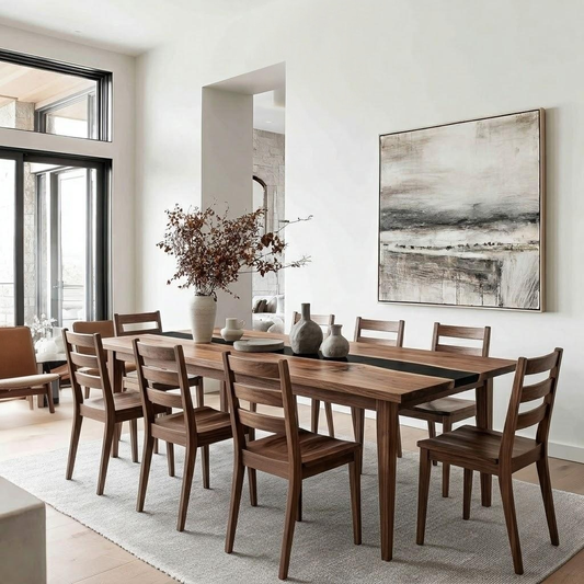 A dining room scene with a rectangular wooden dining table and matching wooden chairs arranged on a light rug. A large abstract black-and-white painting hangs on the wall, and the room is lit by tall windows on the left.