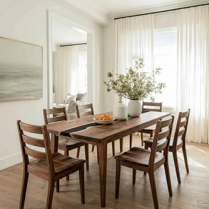A dark wood dining table with six matching ladder-back chairs in a bright, modern dining room. The table is styled with a black runner, a white vase with dried branches, and a bowl of oranges, set against white walls, sheer curtains, and a large mirror.