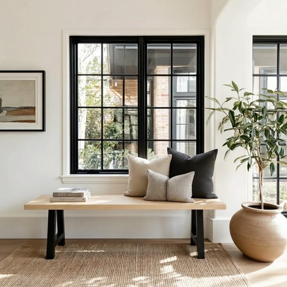 A styled interior scene with a light wood bench against a white wall, set with three throw pillows in beige and charcoal. A large potted plant and a woven rug occupy the foreground, with black-framed windows in the background.