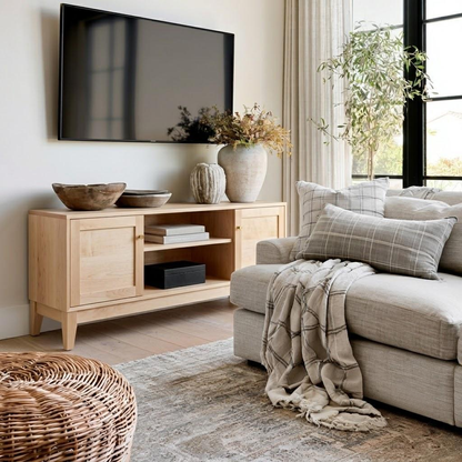 A light natural wood media console in a bright, modern living room setting, styled with a wall-mounted TV, decorative vases, and a throw blanket on a gray sofa.