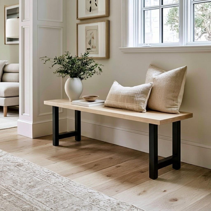 A light natural wood bench with black metal legs in a bright, modern entryway. The bench is styled with two beige throw pillows, a white ceramic vase with greenery, and a small stack of books.
