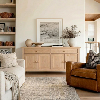 A light natural wood sideboard or credenza in a styled living room, decorated with a framed print, ceramic vases, and a vase of dried branches. The room features a cream sofa, a brown leather armchair, a textured rug, and open shelving on the left.