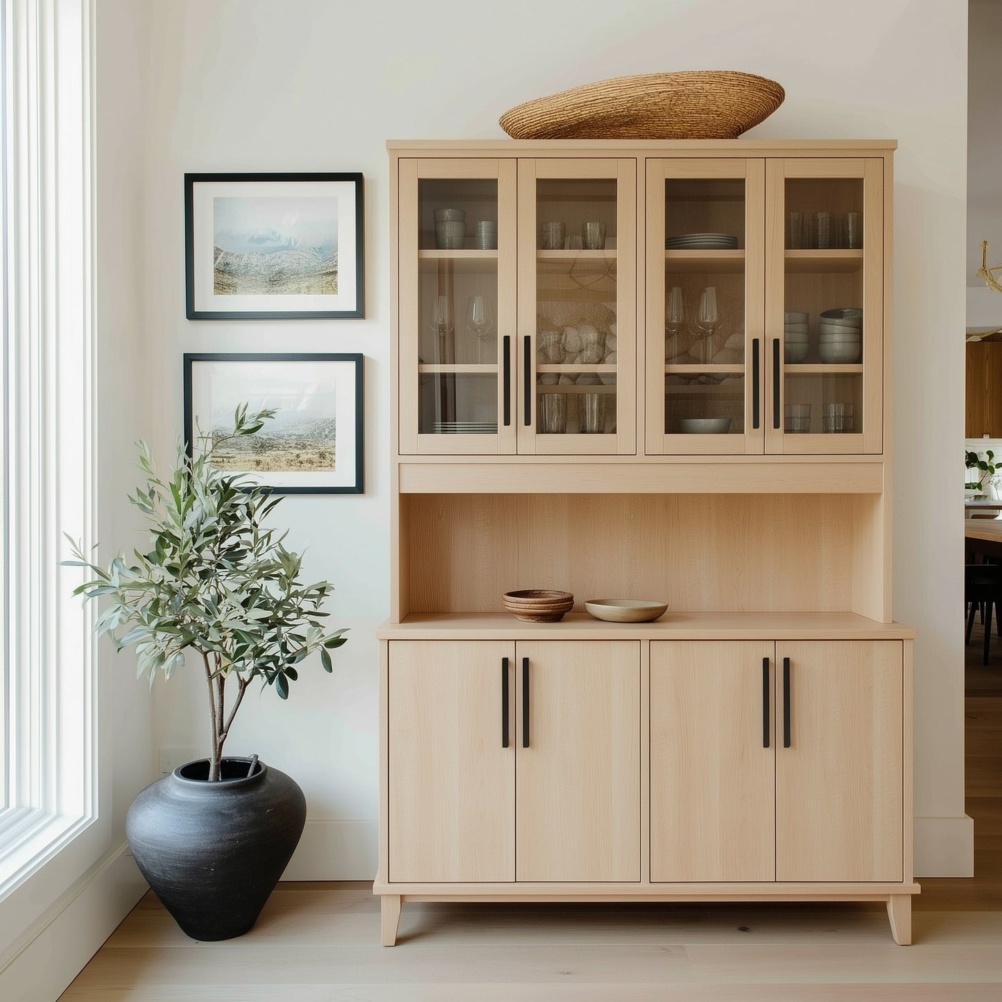 Light wood cabinet with glass doors and dark handles in a room with a vase of plants and framed pictures on the wall.