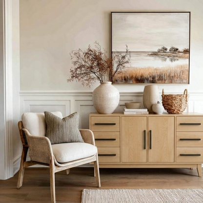 A styled interior scene with a light wood sideboard against a cream wall, decorated with ceramic vases, books, and a woven basket. A matching armchair sits to the left, and a framed landscape painting hangs above the cabinet.