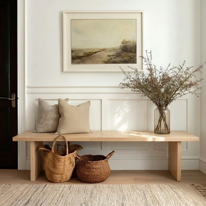 A light natural wood entryway bench styled with two beige pillows, two woven baskets underneath, and a clear glass vase holding dried branches on the right. Above the bench is a framed landscape painting on a white paneled wall.