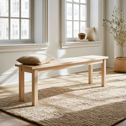A light natural wood bench with a beige cushion, placed on a woven jute rug in a bright, sunlit room with large windows and a potted plant.