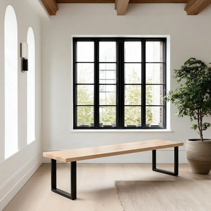 A modern minimalist bench with a light natural wood top and black metal U-shaped legs, positioned in a bright room with a large black-framed window, white walls, and a potted plant.