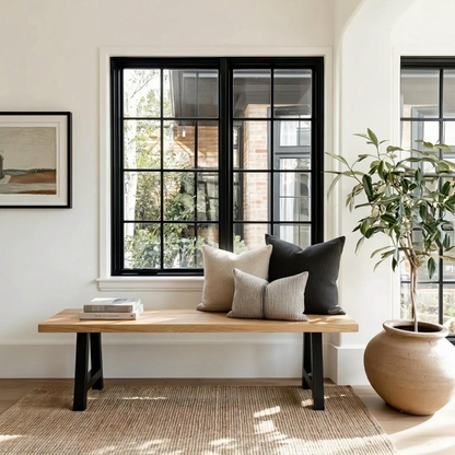 A styled interior scene with a light wood bench against a white wall, decorated with beige and black throw pillows. A large potted plant in a beige ceramic pot stands to the right, and a framed print hangs on the left.