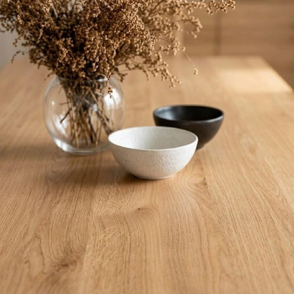 Three ceramic bowls in white, black, and beige tones arranged on a light wood tabletop, with a glass vase of dried flowers in the background.