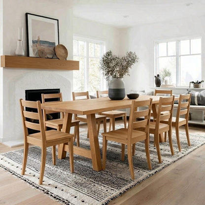 A light natural wood dining table with matching ladder-back chairs, styled in a bright, modern dining room with a fireplace and large windows.