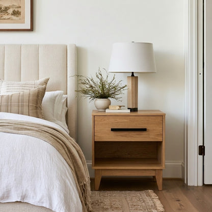 An Oak Astoria Nightstand in a styled bedroom setting, featuring a white drum lampshade lamp, a small ceramic vase with dried branches, and a stack of books on its single drawer front with a black horizontal handle.