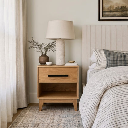 An Oak Astoria Nightstand with a single drawer and open lower shelf, styled in a bedroom next to a bed with striped bedding, a table lamp, and a small vase.