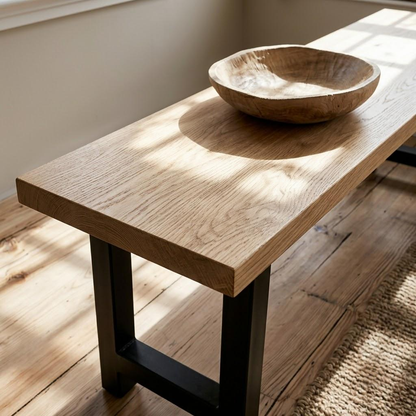 A rustic light oak wood console table with a thick rectangular top and black metal legs, styled with a carved wooden bowl on top in a sunlit room.
