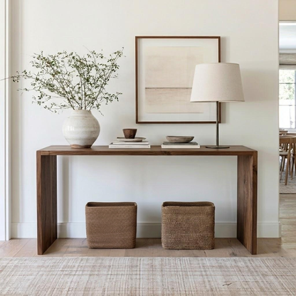 A dark brown wooden console table styled in a bright, modern room with a white vase of greenery, a table lamp, a framed abstract print, and two woven storage baskets underneath.