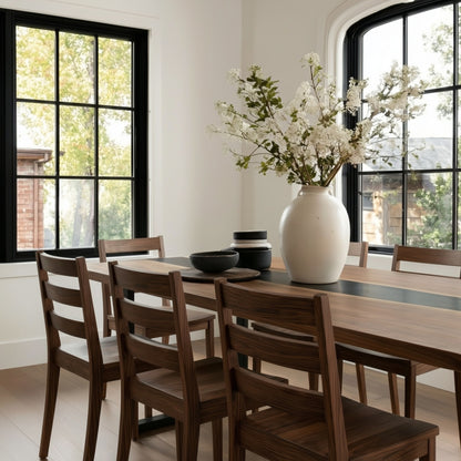 Dining room with wooden table and chairs, large vase with flowers, and large windows.