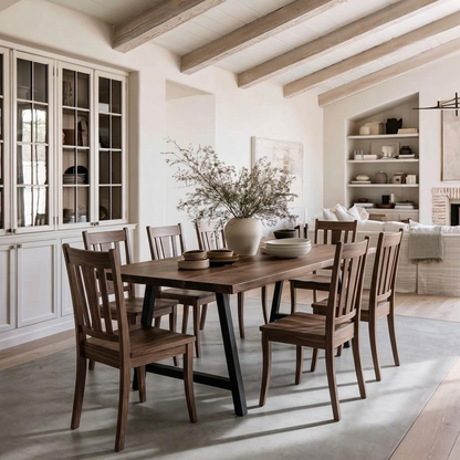 A dining room scene with a rectangular wooden table and six matching wooden chairs, styled with a large vase of dried branches and bowls. The room has exposed wood ceiling beams, white walls, built-in shelving, and a light rug.