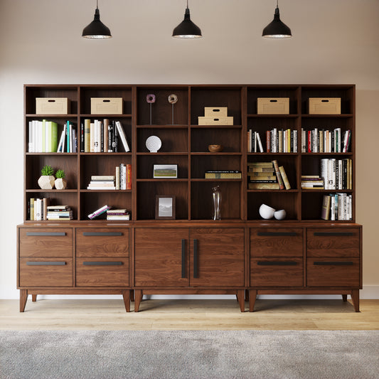 A large wooden sectional bookshelf with multiple shelves and drawers, featuring a dark stain finish, positioned in a room with wall lights above it.
