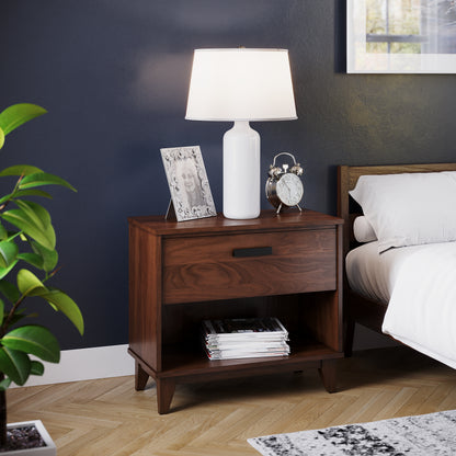 A wooden bedroom nightstand with a lamp and a clock on top, placed next to a bed, with a plant in the background.