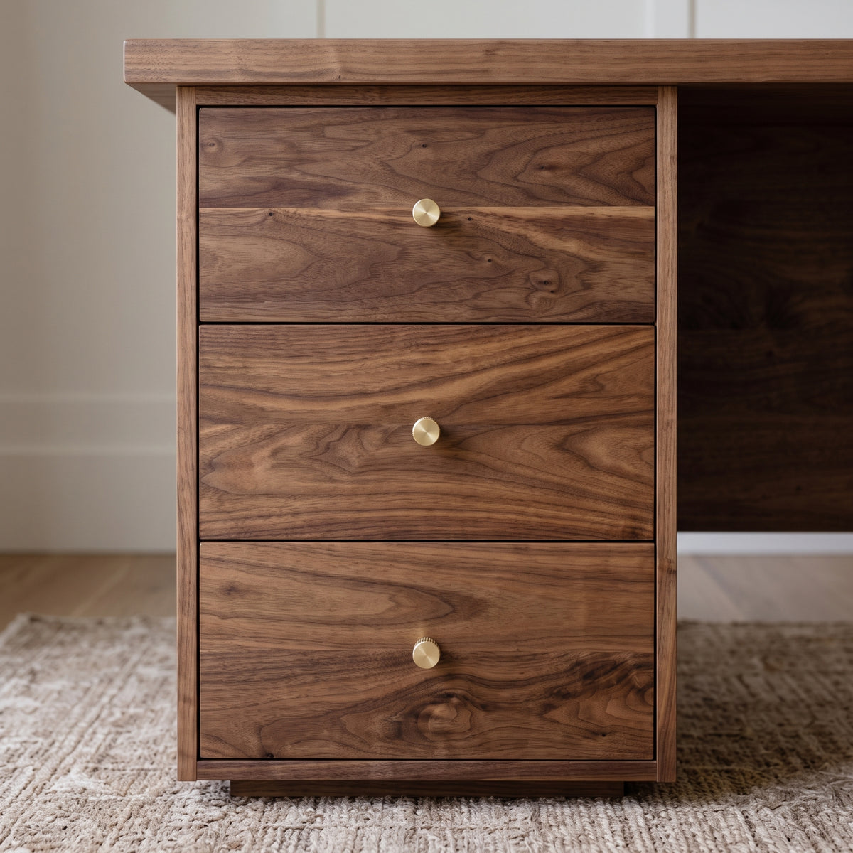 A three-drawer wooden pedestal cabinet with a rich walnut-like grain finish and small round brass knobs, shown in a home office setting on a woven rug.