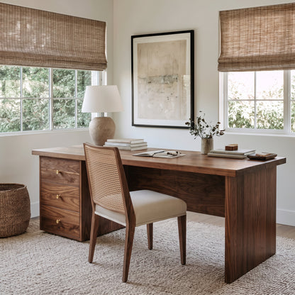 A warm wood writing desk with three drawers and brass handles, paired with a woven-back cane chair in a bright, modern home office. The desk is styled with a ceramic lamp, books, and a small vase, set against white walls with a large framed abstract print.
