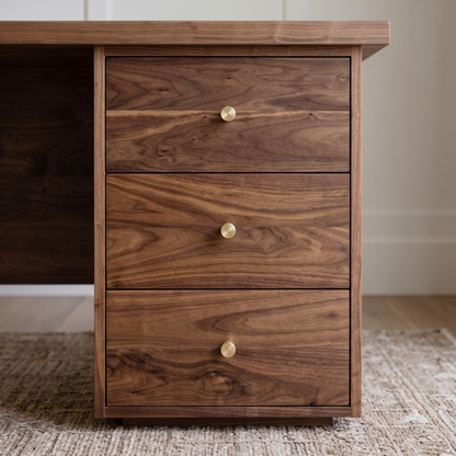 A three-drawer wooden cabinet or desk pedestal with a rich walnut-like wood grain finish and small round brass knobs, shown in a home office setting on a woven rug.