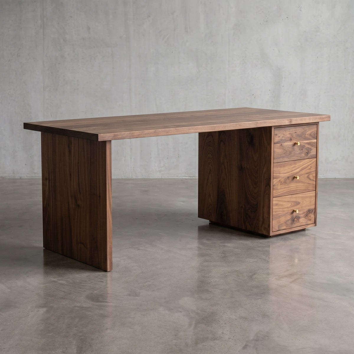 A dark walnut wood writing desk with a solid rectangular top, a right-side pedestal base with three drawers, and brass drawer pulls, set against a concrete wall and floor.