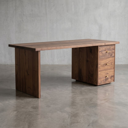 A dark walnut wood writing desk with a solid rectangular top, a right-side pedestal base with three drawers, and brass drawer pulls, set against a concrete wall and floor.