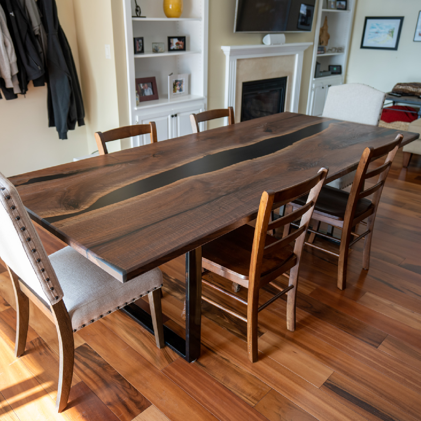 Black walnut dining table with black epoxy river running through the middle in a small dining room with wooden chairs on either side, and a white upholstered chair at the head of the table