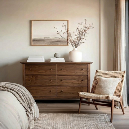 A warm-toned bedroom scene featuring a dark wood dresser with a white vase of dried branches, a framed landscape print, books, and a woven accent chair with a neutral pillow.