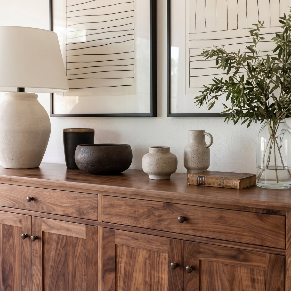 A styled interior scene showing a wooden sideboard or credenza against a white wall, decorated with a ceramic table lamp, framed wall art, ceramic vases, a small book, and a glass vase with greenery.