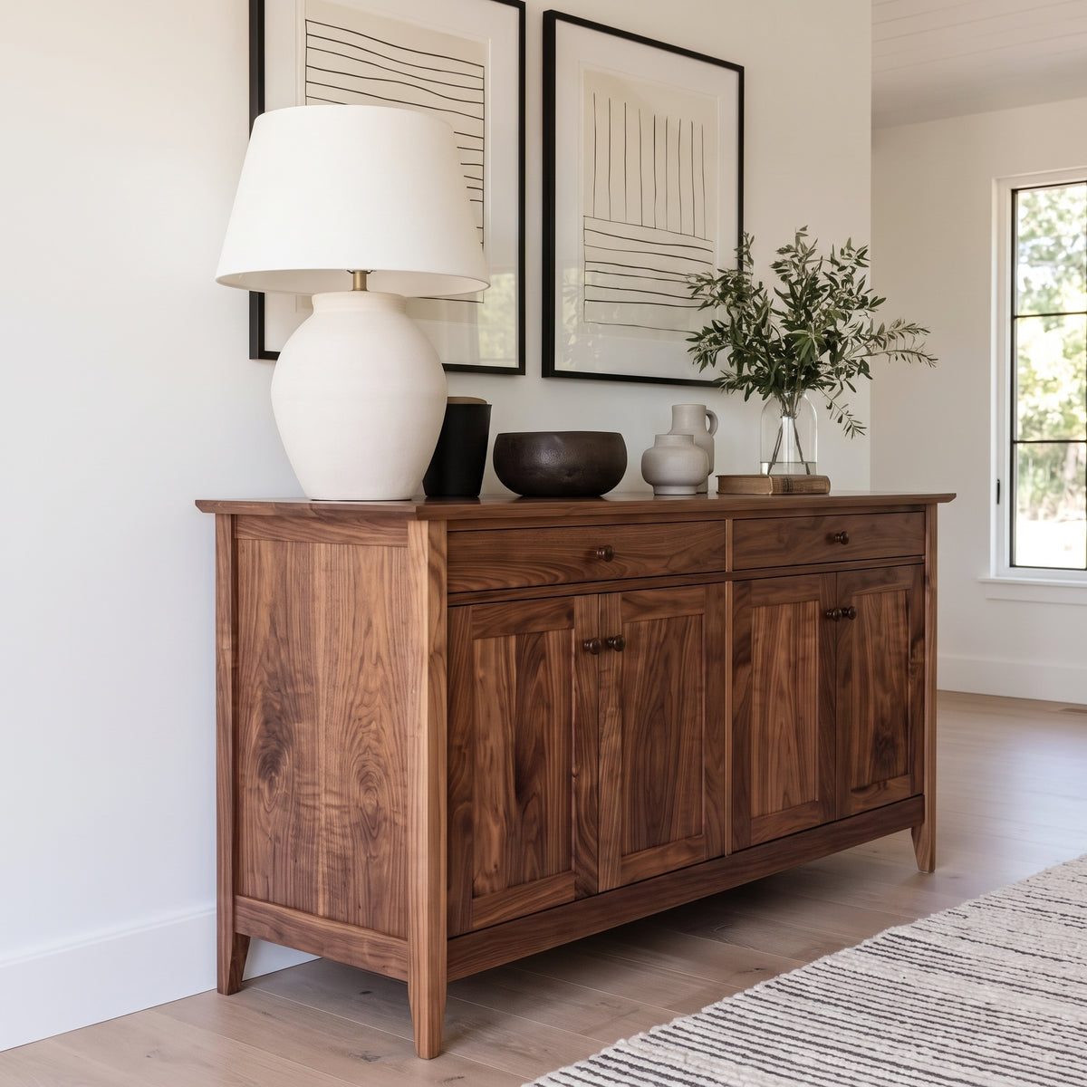 A rustic dark brown wooden sideboard or credenza in a bright, modern room, styled with a white ceramic table lamp, a black bowl, a gray vase, and a glass vase with greenery.