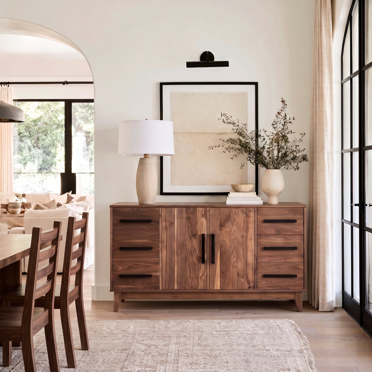 A mid-century modern wooden sideboard cabinet in a warm walnut finish, styled in a dining room with a table, chairs, a lamp, and vases.