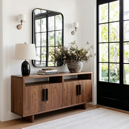 A mid-century modern wooden sideboard cabinet in a warm walnut finish, styled in a bright entryway with a black-framed mirror, wall sconces, a black table lamp, books, and a large ceramic vase with dried branches.
