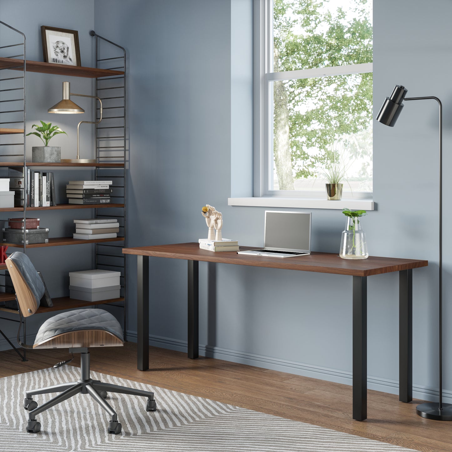 A sleek modern desk with a wooden top and black metal legs, situated in a well-lit room with a chair pulled up to it. Decorative items and a framed picture hang on the wall in the background.