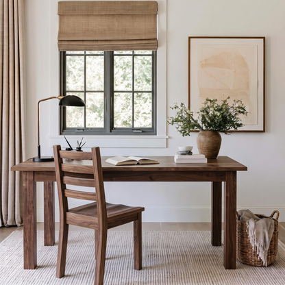 A rustic wooden writing desk with matching ladder-back chair in a bright, neutral-toned home office. The desk is styled with an open book, a black desk lamp, a ceramic vase with greenery, and a stack of books on a light woven rug.