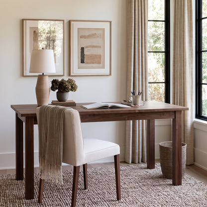 A styled home office scene featuring a dark wood writing desk with a cream upholstered chair, a ceramic table lamp, and two framed abstract art prints on a light beige wall.