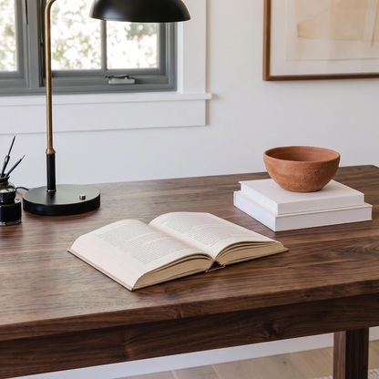A dark wood writing desk in a bright home office setting, styled with an open book, a black desk lamp, a stack of white books, and a terracotta bowl.