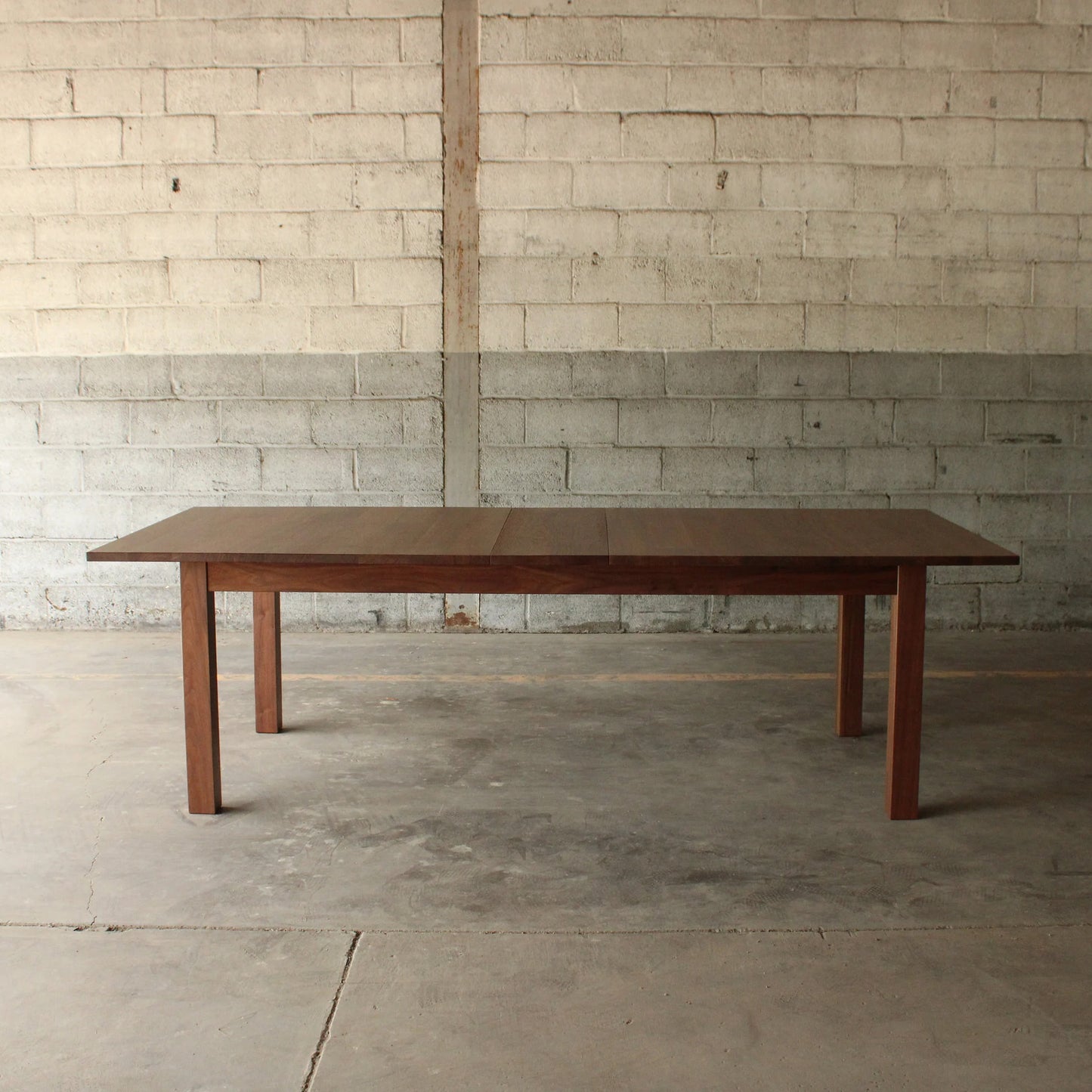 A rectangular dining table with a dark wood finish and metal legs, placed on a concrete floor against a brick wall.