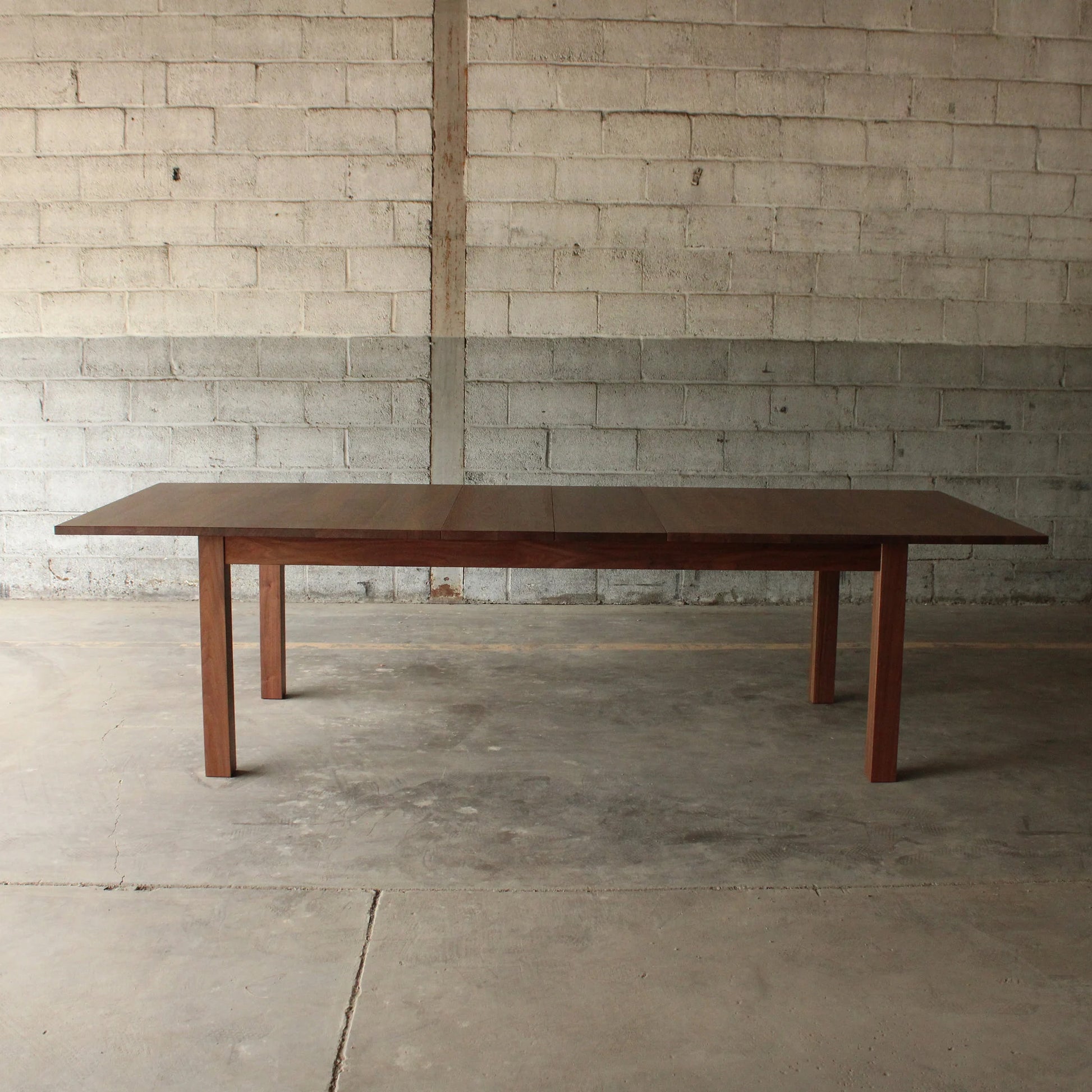 A rectangular dining table with a dark wood finish and metal legs, placed on a concrete floor against a brick wall.