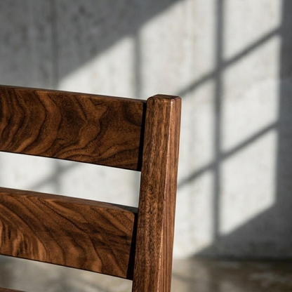 Close-up of a dark brown wooden chair back with rich wood grain, set against a textured gray concrete wall with soft window shadow patterns.