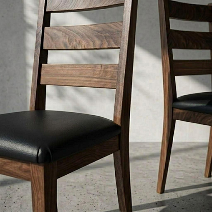 A close-up of a wooden dining chair with a black upholstered seat, shown in a real interior setting with another matching chair partially visible and soft wall shadows.
