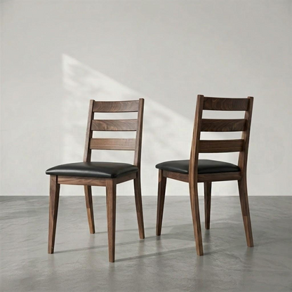 Two wooden dining chairs with black upholstered seats, shown from the back in a minimalist studio setting with a gray floor and light gray wall.