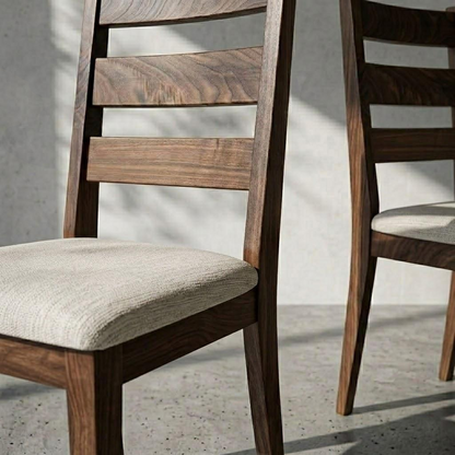 A close-up of a wooden dining chair with a slatted backrest and a light beige upholstered seat, shown in a real interior setting with soft shadows on a concrete floor and wall. Part of a second matching chair is visible at the right edge.