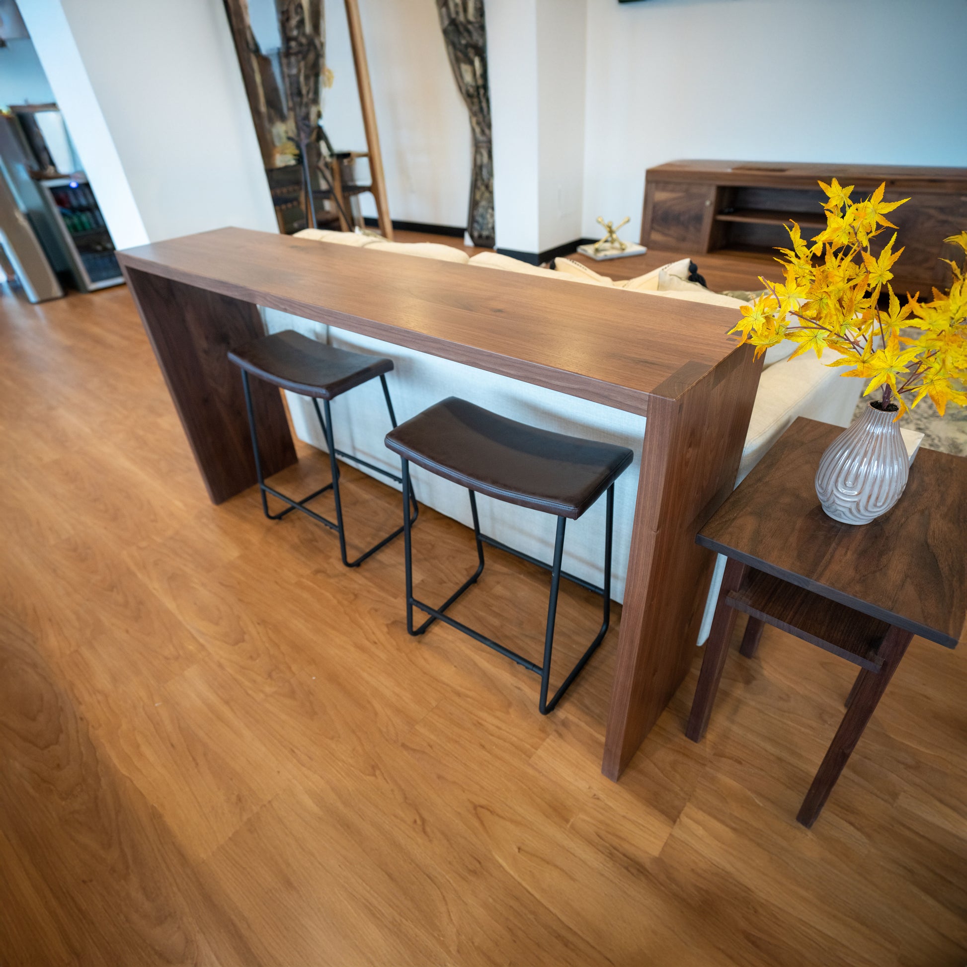 A walnut waterfall sofa table with metal hairpin legs, accompanied by two stools and a vase with yellow flowers, placed on a wooden floor.