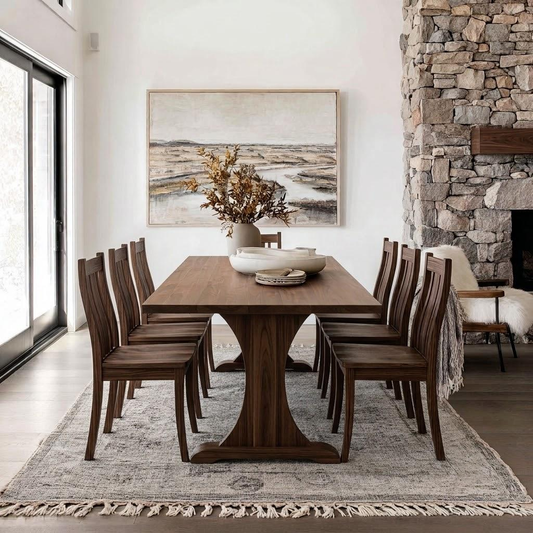 A rectangular dark wood dining table with matching slatted chairs, styled in a bright dining room with a stone fireplace, large window, and framed landscape art on the wall.