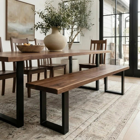 A dining room scene featuring a wooden table with black metal legs, a matching wooden bench, and dark wood chairs, styled with a large beige vase of greenery on a patterned rug.